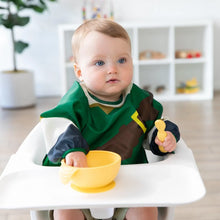 Load image into Gallery viewer, A baby in a high chair holds a yellow bowl and spoon, wearing a Bumkins Sleeved Bib: Zelda™ with shelves and a plant behind.