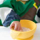 Close-up of a child's hand reaching into a yellow bowl on a high chair tray, wearing a Bumkins Sleeved Bib: Zelda™.