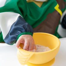 Load image into Gallery viewer, Close-up of a child's hand reaching into a yellow bowl on a high chair tray, wearing a Bumkins Sleeved Bib: Zelda™.
