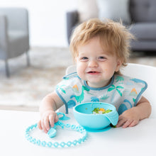 Load image into Gallery viewer, A boy with messy hair smiles, wearing a Bumkins SuperBib® 3 Pack: Hangry, Dinosaurs, Blue Tropic; a blue bowl sits on a white table in front.