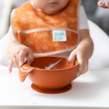 Load image into Gallery viewer, A baby in an orange bib grips a bowl from Bumkins Silicone First Feeding Set: Clay on a high chair tray.