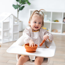 Load image into Gallery viewer, A girl in a beige bib sitting in a high chair smiles as she eats from a Bumkins Silicone First Feeding Set: Clay.