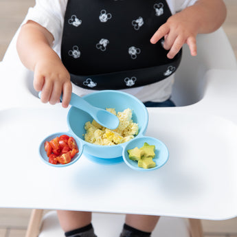 A toddler in a high chair eats scrambled eggs, tomatoes, and avocado from a Bumkins Silicone First Feeding Set: Mickey Mouse Light Blue.