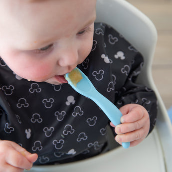A baby in a high chair wears a bib, holding a spoon from Bumkins Silicone First Feeding Set: Mickey Mouse Light Blue.