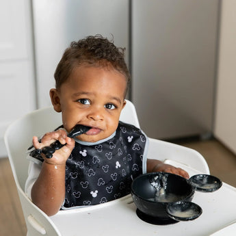 A toddler in a high chair, wearing a black printed bib, eats from Bumkins Silicone First Feeding Set: Mickey Mouse Classic Black.