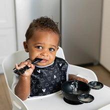 Load image into Gallery viewer, A toddler in a high chair, wearing a black printed bib, eats from Bumkins Silicone First Feeding Set: Mickey Mouse Classic Black.