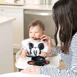 An adult feeds grinning baby in a high chair from a Bumkins Silicone First Feeding Set: Mickey Mouse Classic Black.
