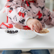 Load image into Gallery viewer, A child in a Minnie Mouse bib reaches for snacks from a Bumkins Silicone First Feeding Set: Mickey Mouse Vanilla Sprinkle on a high chair tray.