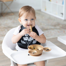 Load image into Gallery viewer, A smiling child wearing a black bib in a high chair eats fruit with Bumkins Silicone First Feeding Set: Minnie Mouse Gold in a bright room.