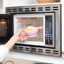 Load image into Gallery viewer, A person places a Bumkins Silicone First Feeding Set: Minnie Mouse Light Pink filled with yellow food into an open microwave.