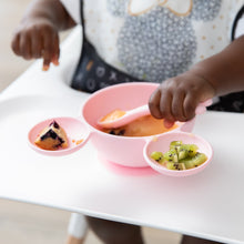 Load image into Gallery viewer, A child in a high chair enjoys using Bumkins Silicone First Feeding Set: Minnie Mouse Light Pink filled with kiwi, applesauce, and a muffin.