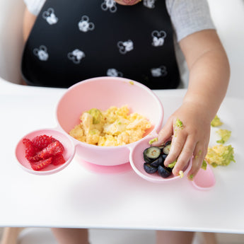 A child in a high chair enjoys scrambled eggs from a Bumkins Silicone First Feeding Set: Minnie Mouse Light Pink, with berries on the side.
