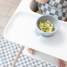 Load image into Gallery viewer, A baby in a checkered bib sits in a high chair, eating from a Bumkins Silicone First Feeding Set: Gray with a checkered mat on the floor.