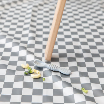 A Bumkins Silicone Dipping Spoons 3 Pack: Taffy lays atop a gray & white checkered mat among pieces of pasta & broccoli by a high chair leg.