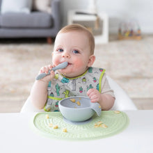 Load image into Gallery viewer, A baby wearing a cactus print bib eats from a Bumkins Silicone First Feeding Set: Gray on a green placemat in a cozy living room.