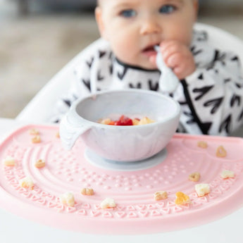 A baby in a black & white bib eats from a Bumkins Silicone First Feeding Set: Marble on a pink silicone mat covered in cereal pieces.