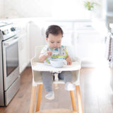 A baby wearing a cactus print bib in a high chair enjoys a meal from a Bumkins Silicone First Feeding Set: Marble in a bright kitchen.