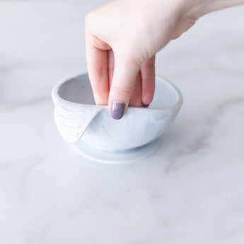 A hand holds a Bumkins Silicone First Feeding Set: Marble, demonstrating the suction cup bottom on a marble surface.