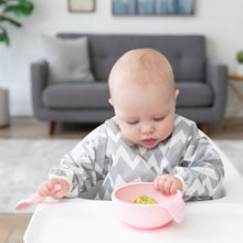 Load image into Gallery viewer, A baby in a gray & white bib eats mashed avocado with Bumkins Silicone First Feeding Set: Pink while seated in a high chair.