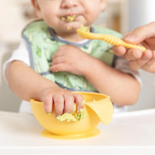 Load image into Gallery viewer, A baby in a green bib in a high chair grabs avocado from a Bumkins Silicone First Feeding Set: Pineapple as a hand holds a spoon nearby.