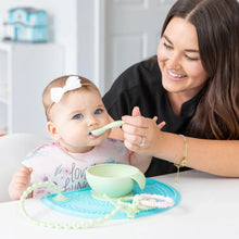 Load image into Gallery viewer, A woman feeds a baby in a floral bib from a Bumkins Silicone First Feeding Set: Sage on a blue silicone placemat next to a teething toy.