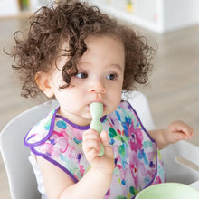 Load image into Gallery viewer, A toddler with curly hair sits at a table, holding a spoon from Bumkins Silicone First Feeding Set: Sage, while wearing a colorful bib.