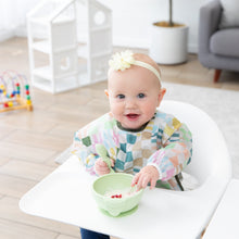 Load image into Gallery viewer, A baby wearing a floral headband and Bumkins Sleeved Bib: Motley Check in a high chair smiles while eating from a green bowl in a playroom.