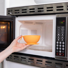 Load image into Gallery viewer, A person places a Bumkins Silicone First Feeding Set: Tangerine into an open microwave.