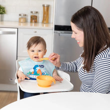 Load image into Gallery viewer, A smiling woman feeds a baby in a high chair with a dinosaur bib from a Bumkins Silicone First Feeding Set: Tangerine.