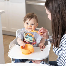 Load image into Gallery viewer, An adult uses an orange Bumkins Silicone Dipping Spoons 3 Pack: Tutti-Frutti to feed a happy baby with an orange bowl in a high chair.