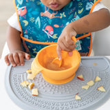 A child in a high chair wearing a blue bib eats from a Bumkins Silicone First Feeding Set: Tangerine on a gray silicone placemat.