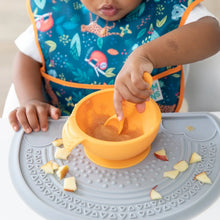 Load image into Gallery viewer, A child in a high chair wearing a blue bib eats from a Bumkins Silicone First Feeding Set: Tangerine on a gray silicone placemat.