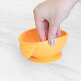 A hand holds a Bumkins Silicone First Feeding Set: Tangerine, demonstrating the suction cup bottom on a marble surface.