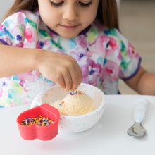Load image into Gallery viewer, A child in a colorful bib adds rainbow sprinkles to ice cream in a Bumkins Silicone First Feeding Set: Vanilla Sprinkle at a white table.