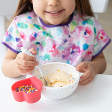 Load image into Gallery viewer, A child in a floral bib beams while enjoying ice cream from a Bumkins Silicone First Feeding Set: Vanilla Sprinkle at a white table.