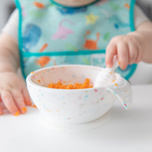Load image into Gallery viewer, Close-up of a baby wearing a colorful bib enjoying orange food from a Bumkins Silicone First Feeding Set: Vanilla Sprinkle at a white table.