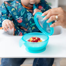Load image into Gallery viewer, A child sits in a high chair chewing a spoon as an adult hand lifts the lid of a Bumkins Silicone First Feeding Set: Blue Jelly with fruit.