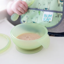 Load image into Gallery viewer, A baby holds a spoon with a Bumkins Silicone First Feeding Set: Green Jelly on a high chair tray while a green bib peeks behind.