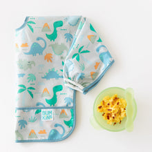 Load image into Gallery viewer, Bumkins Sleeved Bib: Dinosaurs lays folded on a white background next to a green bowl with eggs and a green spoon.