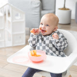 A smiling baby in a gray & white bib messily eats from a Bumkins Silicone First Feeding Set: Pink Jelly while sitting in a high chair.