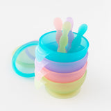 A stack of four colorful bowl and spoon sets, including Bumkins Silicone First Feeding Set: Purple Jelly, on a white background.