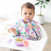 Load image into Gallery viewer, A baby with a colorful bib sits in a high chair using Bumkins Silicone First Feeding Set: Purple Jelly, surrounded by plants and toys.