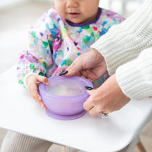 Load image into Gallery viewer, Adult hands guide a toddler with a Bumkins Silicone First Feeding Set: Purple Jelly at a high chair, wearing a colorful bib.