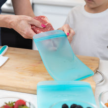 Load image into Gallery viewer, A person puts raw meat in a Bumkins Silicone Flat Reusable Bag 2 Pack, Blue as a child watches at a kitchen counter.