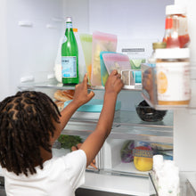 Load image into Gallery viewer, A child places a Bumkins Silicone Flat Reusable Bag 2 Pack, Pink into an open refrigerator next to other bags and a bottle of sparkling water.