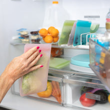 Load image into Gallery viewer, A hand places a Bumkins Silicone Flat Reusable Bag 2 Pack, Pink among lettuce, fruits, and containers into the open fridge.