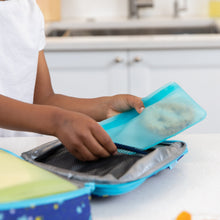 Load image into Gallery viewer, A person packing a Bumkins Silicone Flat Reusable Bag 2 Pack, Blue into an open lunchbox on a kitchen counter.