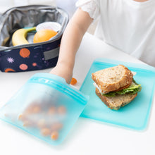 Load image into Gallery viewer, A child's hand reaches for snacks from a Bumkins Silicone Flat Reusable Bag 2 Pack, Blue beside a sandwich, with a lunch bag in the background.