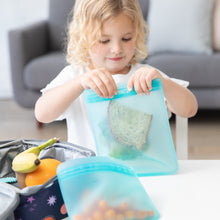 Load image into Gallery viewer, A young child seals a sandwich in a Bumkins Silicone Flat Reusable Bag 2 Pack, Blue at a table next to a lunchbox with fruit.