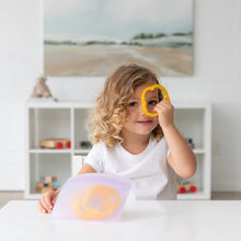 Load image into Gallery viewer, A child peeks through a yellow bell pepper slice at a white table with Bumkins Silicone Flat Reusable Bag 2 Pack, Lavender.